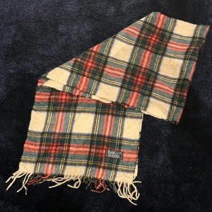 Rutig halsduk från burrberry - Snygg och klassisk rutig halsduk från burrberry i beige, röd, grön och blå. Tillverkad i mjuk ull med fransar i ändarna. Perfekt för att hålla sig varm under kyliga dagar.