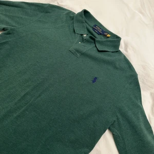 Grön pikétröja från Ralph Lauren - Säljer en klassisk grön pikétröja från Ralph Lauren med broderad logga på bröstet. Tröjan har långa ärmar och en stilren krage med knappar. Perfekt för en avslappnad men stilfull look. Bilden är lite missvisande då tröjan är mer mörkgrön och ser bättre ut i verkligheten då den endast har prövats på. Slimfit Large