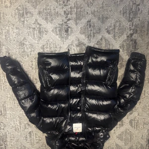 Svart dunjacka från Moncler - Snygg svart dunjacka från Moncler med glansig finish. Jackan har en dragkedja framtill och två fickor med dragkedjor. Perfekt för kyliga dagar med sin varma och puffiga design.