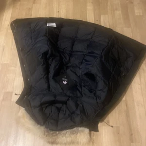 Svart dunjacka med pälsdetalj - Säljer en svart dunjacka från Canada Goose med pälsdetalj på huvan. Jackan har en quiltad design och är perfekt för kalla vinterdagar. Den har en klassisk look med en logotyp på ärmen.