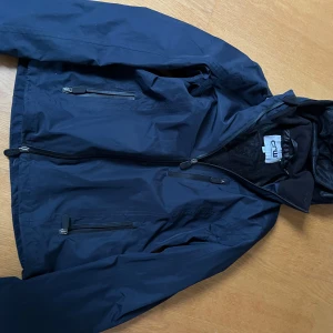 Mörkblå vindjacka från CRW - Säljer en mörkblå vind och ridjacka från CRW i storlek XS. Jackan har dragkedja framtill och två praktiska fickor med dragkedjor. Perfekt för blåsiga dagar och har en stilren design.