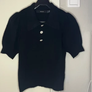 Tröja från VERO MODA - Snygg svart tröja från VERO MODA med tre dekorativa silvriga knappar framtill. Tröjan har korta ärmar och en klassisk krage, perfekt för en stilren look.💯