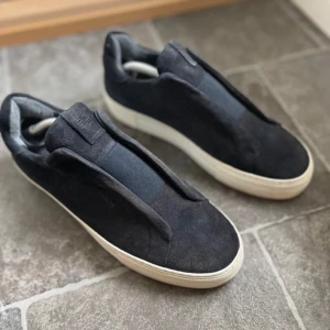 Axel arigato - Snygga svarta sneakers från Axel Arigato i mocka med en stilren design. Skorna har en vit sula och saknar traditionell snörning för en minimalistisk look.