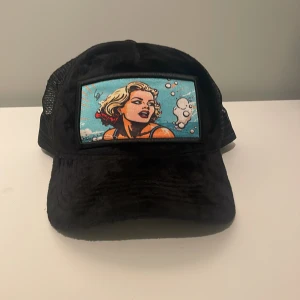 Svart revenite keps som inte säljs längre -  svart keps från Revenite med ett färgglatt popkonstmotiv framtill. Kepsen har ett nätmaterial på baksidan för bättre ventilation och justerbar passform. Perfekt för en avslappnad stil.