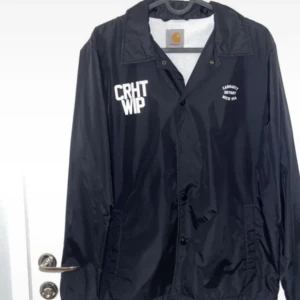Svart coachjacka från Carhartt WIP - Snygg svart coachjacka från Carhartt WIP med tryck på bröstet och ryggen. Jackan har knappar framtill och en klassisk krage. Perfekt för en stilren look.