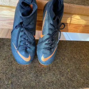 Säljer ett par svarta och orangea Nike Mercurial fotbollsskor. De har en snygg design med snörning och en hög krage för extra stöd. Skorna har dobbar som passar perfekt för spel på gräsplaner. De är i bra skick och redo för nästa match!