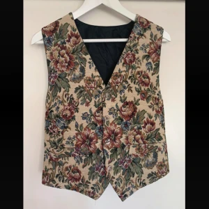 Blommig vintage väst - Väst i blommigt mönster med knappar framtill. Den är köpt på second hand och saknar märke. Det står storlek L men har justerbar baksida och passar därför fler storlekar. Endast använd några fåtal gånger.