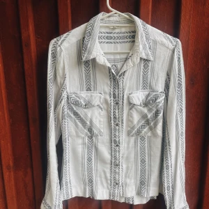 Mönstrad skjorta från River Island - Säljer en superfin skjorta från River Island i storlek 12. Den är vit med ett snyggt svart mönster som ger en bohemisk känsla. Skjortan har långa ärmar och två bröstfickor med knappar. Perfekt för både vardag och fest!