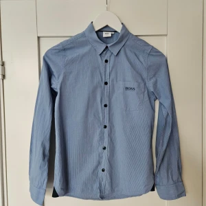 Blå randig skjorta från Hugo Boss - Säljer en stilren blå randig skjorta från Hugo Boss i storlek XS. Skjortan har en slim fit passform och är långärmad med svarta knappar. Perfekt för både vardag och fest!