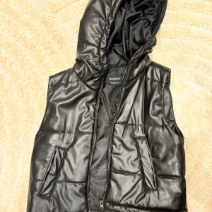 Svart quiltad väst med huva från Busem - Säljer en snygg svart quiltad väst från Busem, är i storlek S men passar likaväl XS. Den har en praktisk huva och dragkedja framtill. Perfekt för höst och vinter när du vill hålla dig varm och stilren. Västen är i bra skick och redo för nya äventyr!