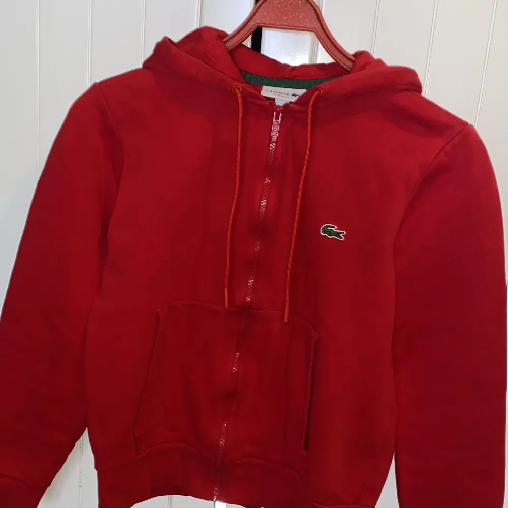 Säljer en snygg röd hoodie från Lacoste i storlek XS. Den har en klassisk passform med dragkedja och den ikoniska krokodilloggan på bröstet. Perfekt för höst och vår, och den är i nyskick. Superbekväm och stilren!. Tröjor & Koftor.