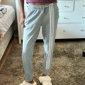 Adidas träningsbyxa  - Grå adidas träningsbyxa med klassiska vita ränder längs sidorna. Medelhög midja med dragsko för justerbar passform. Mudd i bensluten för en smalare silhuett. Nästan oanvänd och i mycket gott skick. Nypris: 500kr. Använd gärna ”köp nu”