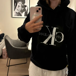 Svart hoodie från Calvin Klein - Säljer en snygg och bekväm svart hoodie från Calvin Klein. Den har en stor logga på framsidan och är perfekt för en casual look. Hoodien har långa ärmar och en klassisk känguruficka. Perfekt för höst och vinter!