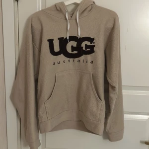 Ugg fleece hoodie - Tja säljer denna mysiga beige hoodie från UGG i fleece-material. Perfekt för kyliga dagar. Den har en stor ficka fram och justerbar huva med vita snören. Hör av vid funderingar!