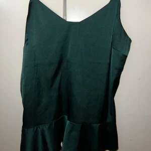 Mörkgrön fest linne - Snygg mörkgrön fest topp i ett mjukt och bekvämt material. Perfekt för både vardag och festliga tillfällen. Den har en enkel design med en liten volangdetalj längst ner som ger en extra touch. Passar bra till jeans eller kjol för en stilren look.