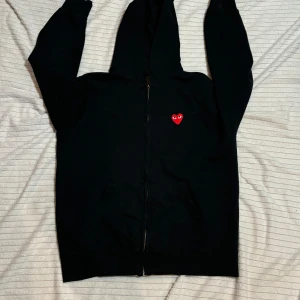 Svart hoodie från Comme des Garçons - Säljer en svart hoodie från Comme des Garçons i storlek Xl men passar nog även L.med det ikoniska röda hjärtat broderat på bröstet. Den har en dragkedja framtill och är i mycket bra skick. Perfekt för höst och vår, och passar till både vardag och fest. Storleken är inte angiven men den ser ut att ha en normal passform.