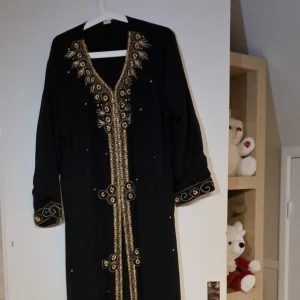 Svart ABAYA med gulddetaljer - Elegant svart lång Abaya från dubai med vackra gulddetaljer och broderier. Klänningen har långa ärmar och en unik design med dekorativa mönster längs framsidan och ärmsluten. Perfekt för festliga tillfällen där du vill sticka ut med stil.