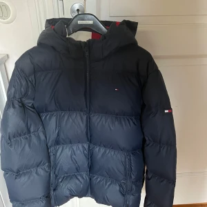 Blå dunjacka från Tommy Hilfiger - Säljer en snygg blå dunjacka från Tommy Hilfiger i storlek 176. Jackan har en cool röd insida och en praktisk huva. Perfekt för kalla vinterdagar med sin varma och puffiga design. Den har en dragkedja framtill och är i mycket bra skick. Passar både till vardags och lite finare tillfällen.