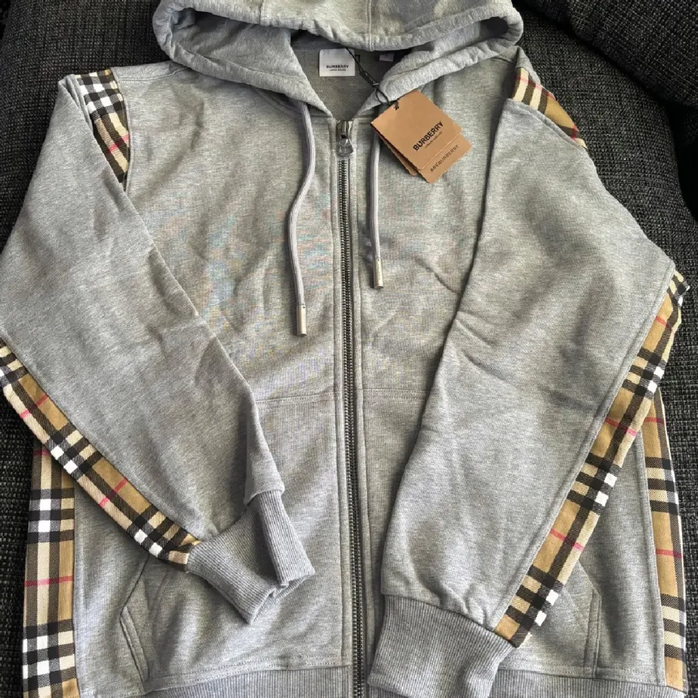 Säljer en snygg grå hoodie från Burberry i nytt skick. Den har en dragkedja framtill och klassiska Burberry-rutor längs ärmarna. Perfekt för höst och vår. Ett måste för garderoben! Pris går att diskuteras!. Tröjor & Koftor.