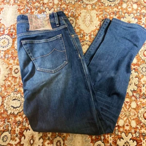 Blå jeans från Jacob Cohen - Säljer ett par snygga blå jeans från Jacob Cohen. De har en klassisk femficksdesign och knappar i gylfen. Jeansen är i bra skick och passar perfekt för både vardag och fest. De har en normal passform och är tillverkade i högkvalitativt denim.