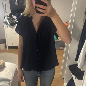 Svart blus med knappar - Säljer en stilren svart blus med knappar framtill. Den har en avslappnad passform och korta ärmar, perfekt för både vardag och fest. Blusen är i mycket bra skick och passar till de flesta outfits.