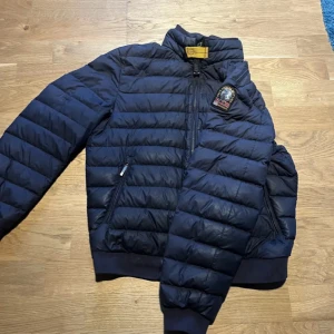 Mörkblå pufferjacka - Säljer en snygg mörkblå pufferjacka i mycket bra skick. Jackan har en dragkedja framtill och är långärmad med ribbade muddar. Perfekt för höst och vinter, håller dig varm och stilren. Den har en broderad detalj på bröstet som ger en extra touch.
