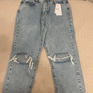 Blå mom jeans från Gina Tricot - Säljer ett par snygga ljusblå mom jeans från Gina Tricot i storlek 40. De har en cool slitning vid knäna och är i nytt skick med etikett kvar. Perfekta för en avslappnad stil!