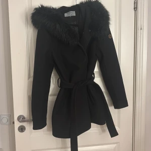 Svart kappa med pälsdetaljer från Hollies - Snygg svart kappa från Hollies med pälsdetaljer på huvan. Den har ett bälte i midjan för en elegant passform och är perfekt för kyliga dagar. Kappan är långärmad och har en stilren design som passar både till vardags och fest.