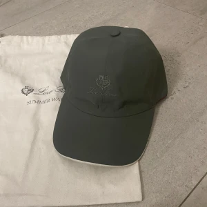 Grön keps från Loro Piana - Snygg grön keps från Loro Piana i ett stilrent och klassiskt utförande. Kepsen har en broderad logga framtill och ett rutigt mönster på insidan. Perfekt för sommardagar och ger en elegant touch till din outfit.