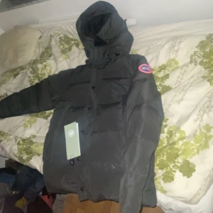 Svart dunjacka från Canada Goose - Säljer en svart  jacka från Canada Goose i oanvändt skick (helt ny). Jackan har en pufferstil med långärmade ärmar och en praktisk huva. Perfekt för kalla vinterdagar. Den har både dragkedja och knappar för extra skydd mot kylan. Märkets ikoniska logga syns på ärmen. Storlek S (173-179cm) passar utmärkt. 