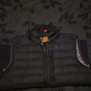 Svart dunväst från Parajumpers - Säljer en svart dunväst från Parajumpers i mycket bra skick. Västen har en dragkedja framtill och en hög krage med en gul detalj. Perfekt för höst och vinter, håller dig varm och stilren. Passar både till vardags och lite mer uppklädda tillfällen.