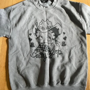 Betty Boop sweatshirt - Säljer en grå sweatshirt med ett coolt Betty Boop-tryck i cowboy-stil. Tröjan är i bra skick och har korta ärmar. Perfekt för en avslappnad look eller som statement piece i garderoben. 🖤Den är S men är oxå baggy!