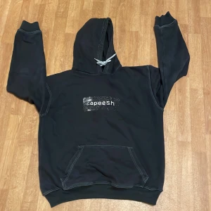 Svart hoodie från Capeesh - Säljer en svart hoodie från Capeesh, perfekt för höst och vinter. Den har en stor ficka fram och en justerbar huva med vita snören. Trycket på framsidan är lite slitet men ger en cool vintage-look. Perfekt för en avslappnad stil!