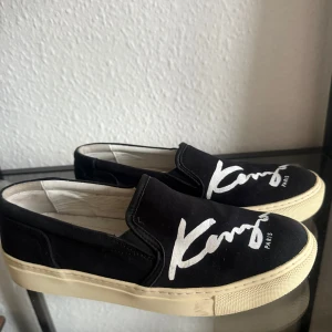 Svarta slip-on sneakers från Kenzo - Säljer ett par svarta slip-on sneakers från Kenzo i bra skick. De har en vit sula och Kenzo-loggan i vitt på ovansidan. Perfekta för en avslappnad stil och lätta att matcha med olika outfits. Skorna är bekväma och stilrena, idealiska för både vardag och fest.