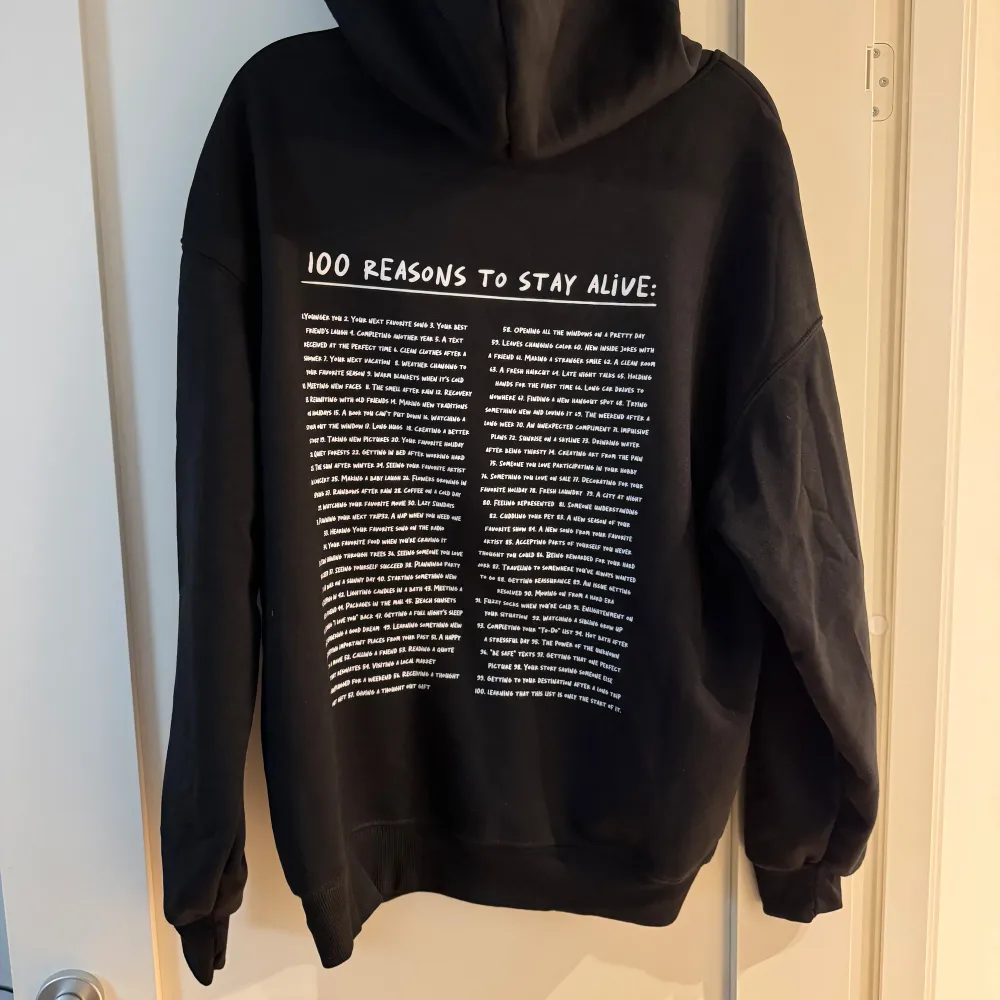 Säljer en svart hoodie med texten 'KEEP GOING:' på framsidan och '100 REASONS TO STAY ALIVE:' på baksidan. Den har en känguruficka och justerbar huva med snören. Perfekt för att hålla sig varm och motiverad under kyliga dagar. Passar både för vardag och mysiga hemmakvällar.. Tröjor & Koftor.