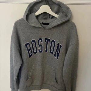 Grå hoodie med Boston-tryck - Säljer en grå hoodie med stort Boston-tryck i mörkblått på framsidan. Den är supermjuk och perfekt för kyliga dagar. Har en stor ficka fram och en mysig huva. Perfekt för en avslappnad stil eller en dag på stan. 🏙️
