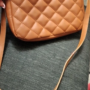 Brun quiltad axelväska - Säljer en snygg brun axelväska i quiltat mönster. Perfekt för vardagsbruk med en justerbar rem. Väskan är i mycket bra skick och har en praktisk dragkedja för säker förvaring. Passar till både höst och vår outfits!