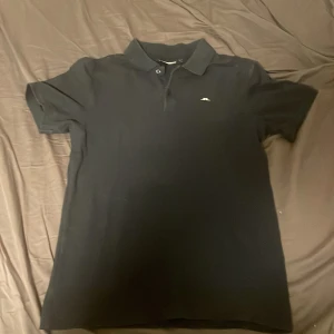 Blå pikétröja från J.Lindeberg - Säljer en stilren svart pikétröja från J.Lindeberg i storlek S. Tröjan har en klassisk krage med två knappar och en liten logga på bröstet. Perfekt för både vardag och festliga tillfällen.