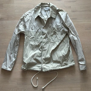 Beige overshirt/jacka Weekday - Säljer en beige med hint av grönt overshirt/jacka från Weekday i bra skick. Den har långa ärmar och knappar framtill. Justerbar dragsko i nederkant för en anpassad passform. Skön och stilren för en casual look.