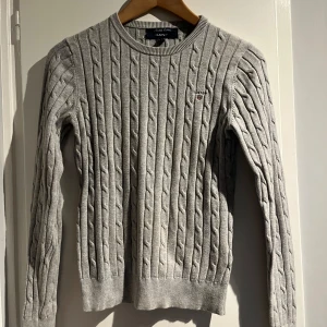 Grå stickad tröja från GANT - Säljer en snygg grå stickad tröja från GANT. Den har ett klassiskt flätmönster och är perfekt för både höst och vinter. Tröjan är långärmad och har en rund halsringning. Perfekt för att hålla sig varm och stilren! 🧶