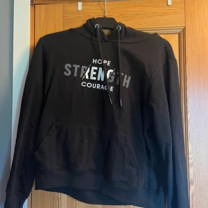 Svart hoodie från OTP - Säljer en svart hoodie från OTP med texten 'Hope Strength Courage' i vitt och grått på framsidan. Den har en klassisk känguruficka och justerbar huva med snören. Perfekt för en avslappnad stil eller mysiga dagar hemma. Endast använd ett par gånger ☺️