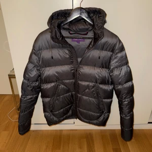 Mackay Down Jacket Ralph Lauren Purple Label - Säljer en snygg svart pufferjacka från Ralph Lauren i storlek S. Jackan har en praktisk huva och dragkedja framtill. Perfekt för kalla dagar med sin varma och quiltade design. Den är i mycket bra skick och redo för nya äventyr! 🖤
