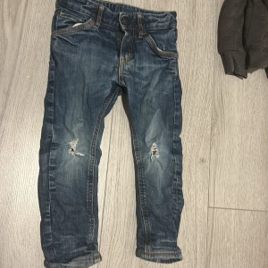 Blå jeans för barn från H&M - Säljer ett par coola blå jeans för barn från H&M i storlek 98. De har en snygg sliten look med hål på knäna och är i tapered stil. Perfekta för vardagsbruk och ger en trendig touch till vilken outfit som helst. De är i bra skick och redo för nya äventyr!