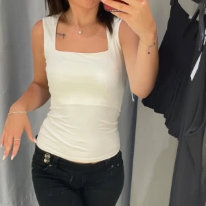Vit ärmlös topp - Säljer en stilren vit ärmlös topp i ett mjukt och stretchigt material. Perfekt för sommardagar eller som ett basplagg i garderoben. Toppen har en enkel och elegant design som passar till både jeans och kjol.