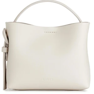 Vit handväska från Arket - Säljer en stilren vit handväska från Arket i skinn. Väskan har en minimalistisk design med en elegant knytdetalj på sidan. Perfekt för både vardag och fest. Den är i mycket bra skick och redo för nya äventyr!