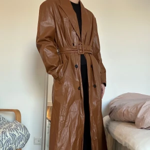 Brun trenchcoat med bälte - Säljer en snygg brun trenchcoat i syntetmaterial. Den har en klassisk design med knappar och ett bälte i midjan för en stilren look. Perfekt för höst och vår, och ger en elegant touch till vilken outfit som helst. Jackan har långa ärmar och en skön passform.