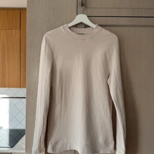Beige tröja från COS - Säljer en stilren och enkel beige tröja från COS. Den är gjord i 100% bomull vilket gör den superbekväm och perfekt för vardagsbruk. Tröjan har långa ärmar och en ribbad krage som ger en snygg detalj. Perfekt för både höst och vår!