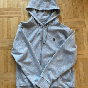 Grå hoodie med dragkedja - Säljer en snygg och bekväm grå hoodie från Polo Ralph Lauren. Den har en dragkedja framtill och en klassisk logga på bröstet. Perfekt för både vardag och chill. Storleken är herr M och den är i mycket bra skick. Passar perfekt till höst och vår!