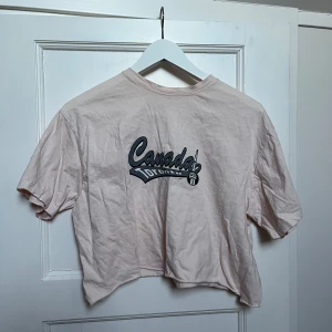 Rosa croppad t-shirt med tryck - Säljer en rosa croppad t-shirt med ett coolt 'Canada' tryck på framsidan. Perfekt för en avslappnad stil. T-shirten är kortärmad och i bra skick. Passar perfekt till jeans eller shorts för en chill look.
