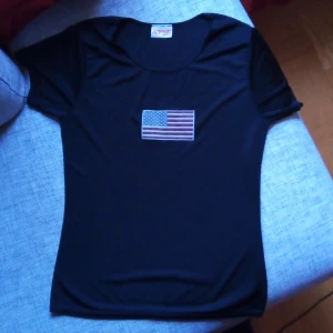 Svart t-shirt med amerikansk flagga från Revival - Säljer en svart t-shirt från Revival Girlswear med en amerikansk flagga tryckt på framsidan. T-shirten är kortärmad och i bra skick, perfekt för en avslappnad stil. Passar till både jeans och shorts!